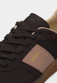 Gros plan d'une chaussure de sport noire et beige avec des lacets noirs, un logo "BOSS" doré sur le côté beige, et une doublure intérieure beige.