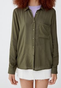 Chemise olive à manches longues avec boutons, en tissu lisse, dotée d'une poche poitrine. Portée par-dessus un haut violet clair, associée à une jupe blanche à ourlet effiloché.