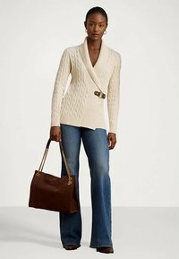 Mulher vestindo um suéter creme de malha tipo cabo com cinto, jeans azuis, botas castanhas, carregando uma grande bolsa castanha com alças de corrente douradas.