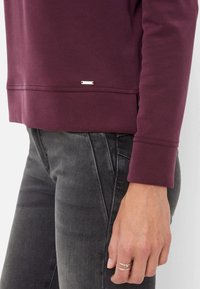 Sweatshirt bordeaux à manches longues, avec un logo argenté près de l'ourlet et des poignets côtelés, associé à un jean gris foncé.