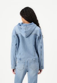 Svetlo modra denim jakna s kapuco, z udobnim krojem, vidnimi šivi in stranskimi žepi. Nosimo jo z ujemajočimi svetlo modrimi kavbojkami.