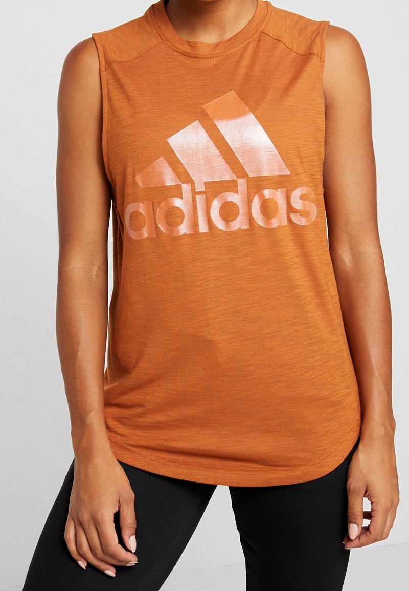 Personne portant un débardeur orange Adidas avec un grand logo, associé à des leggings noirs, debout devant un fond gris uni.