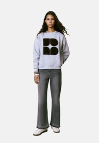 Sudadera gris con diseño gráfico negro, puños acanalados, combinada con pantalones acampanados grises y zapatillas blancas. El fondo es de color blanco liso.