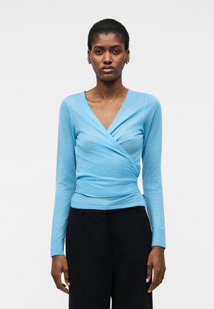 Žena stojaca v svetlomodrom zavinovacom svetri s dlhým rukávom a čiernych vysokých nohaviciach na bielom jednoduchom pozadí.