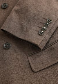 Manche et partie avant d'un blazer marron texturé montrant quatre boutons de manche, un bouton de devant et une poche à rabat.