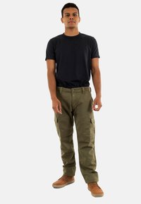 Olijfgroene cargobroek met meerdere zakken, gecombineerd met een getailleerd zwart T-shirt en bruine veterschoenen. Model staat rechtop.