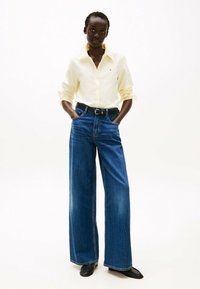 Camisa amarilla de botones con las mangas remangadas, combinada con unos vaqueros de pierna ancha de denim azul y un cinturón negro, completada con zapatos negros.