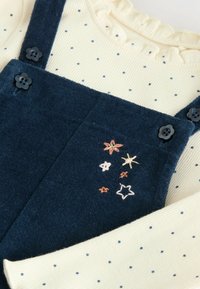 Marineblauwe corduroy tuinbroek met bloemen- en sterrenborduurwerk, gecombineerd met een romige geribbelde top met kleine marineblauwe stippen. Zwarte knoopsluitingen.