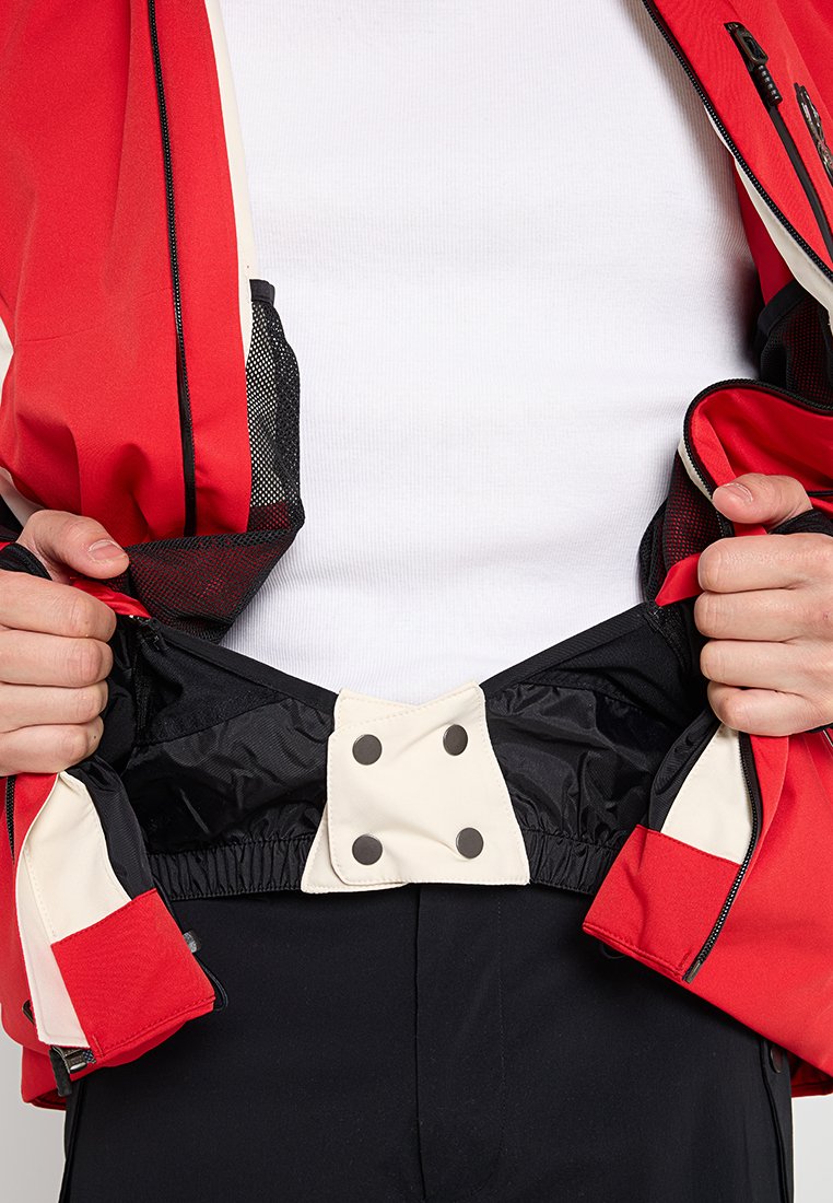 Rote und schwarze Jacke mit einem cremefarbenen Innenfutter, die einen Reißverschluss und Druckknöpfe an der Taille aufweist. Texturiertes Material ist sichtbar.