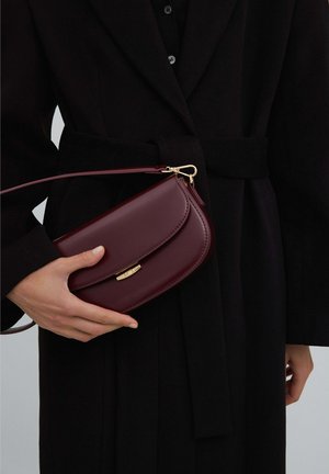 Persona che indossa un cappotto nero e tiene una piccola borsa in pelle bordeaux con chiusura dorata e tracolla staccabile.