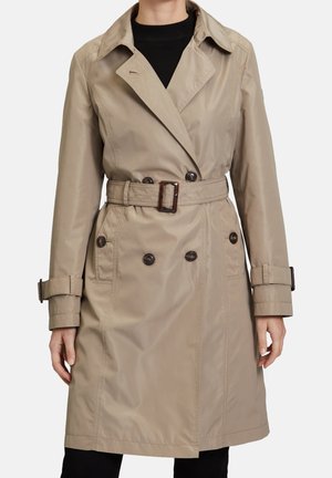 Femme portant un trench-coat double boutonnage beige avec ceinture et poignets boutonnés par-dessus un haut noir.