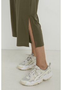 Ben klädda i olivgröna kapade byxor med sidoöppningar, kombinerat med vita och beige snörade sportskor på ett ljust golv.