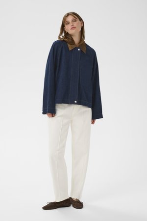 Woman wearing a dark blue jacket with a brown collar, white straight-leg pants, and dark brown flat shoes, standing against a plain background.