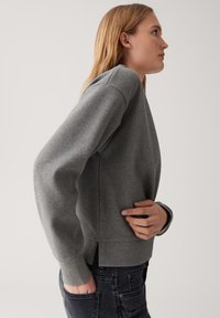 Donna in un maglione grigio a maniche lunghe con spacco laterale, jeans neri, una mano in tasca, rivolta a destra su uno sfondo chiaro uniforme.