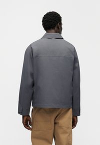 Γκρι μπουφάν με γιακά, μακριά μανίκια και λεπτομέρειες με κουμπιά. Στο πίσω μέρος έχει οριζόντια ραφή. Φορεμένο με μπεζ παντελόνι τύπου cargo.