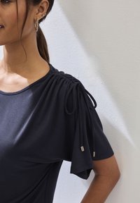 Camisa de marina con detalle de hombro fruncido, que presenta lazos con acentos metálicos. Suave tejido y corte holgado mejoran la comodidad.