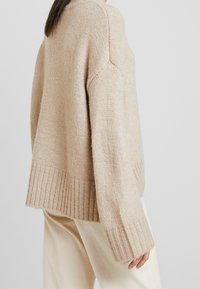 Pull en maille beige avec une coupe ample, poignets et ourlet côtelés, caractérisé par une texture douce et une couture d'épaule tombante. Associé à un pantalon de couleur claire.