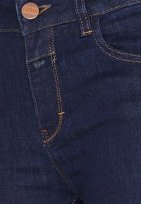 Gros plan sur un jean en denim bleu foncé avec des coutures orange, un bouton en cuivre, un passant de ceinture et une petite étiquette près de la poche indiquant "Closed."