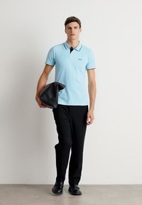 Polo bleu clair avec des accents de col noir, associé à un pantalon noir. Le mannequin tient un sac noir, marchant devant un fond uni.