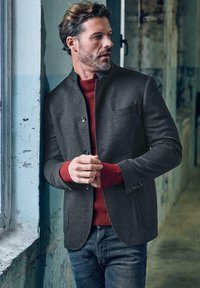 Grey tailored jacket with a high collar, featuring a chest pocket and buttons, layered over a red ribbed sweater, worn with blue jeans.