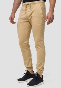 Beige joggerbukser med elastisk talje, frontlommer og tapered ankler. Styles med sorte sneakers med hvide såler.