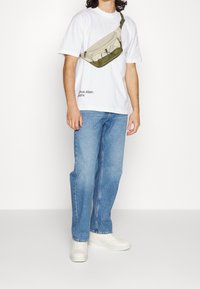 Beżowa i oliwkowo-zielona torba na ramię z zamkiem błyskawicznym, noszona na białej koszulce i niebieskich dżinsach; w połączeniu z białymi trampkami.