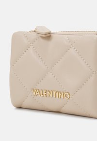 Valentino bež kožna torbica s odabranim uzorkom, koja sadrži zlatne detalje s logotipom i zatvaranje na zip. Teksturna, glatka površina.