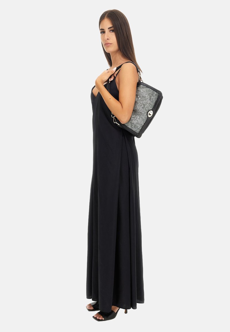 Abito lungo nero con spalline sottili, caratterizzato da un design fluido. La modella porta una borsa a tracolla grigio metallico con un bottone in metallo argentato.
