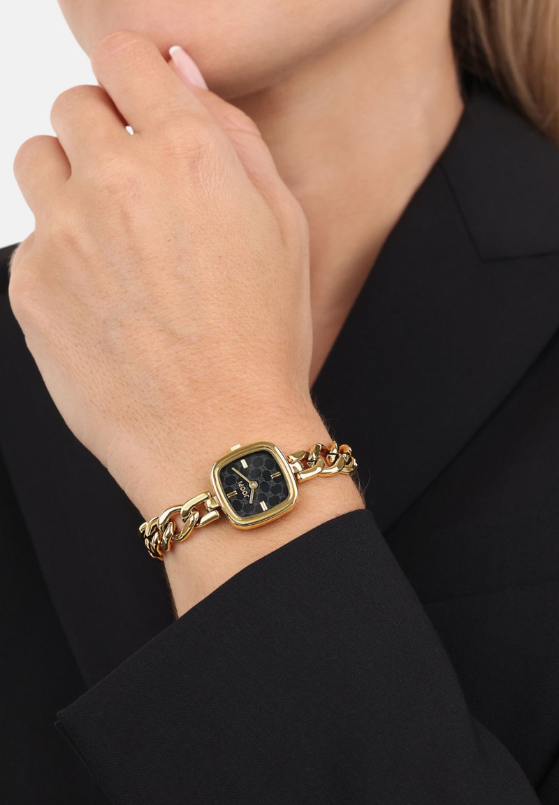 Goldfarbene Armbanduhr mit quadratischem Zifferblatt, schwarzem hexagonalem Muster und einem verbundenen Armband. Schwarzer Hintergrund mit sichtbarem Hautton.