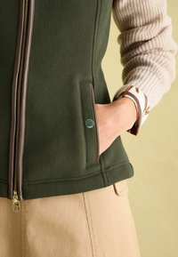 Grüne Fleece-Weste mit einem braunen Reißverschluss und Taschen, über einem beigen Strickärmel mit einem gemusterten Bündchen getragen. Einfaches Design und strukturierte Oberfläche.