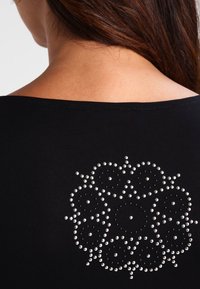 Svart topp med rund halsringning och en bakdekoration av silverfärgad nitar arrangerade i ett blommönster mot en enfärgad bakgrund.