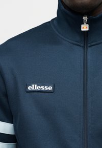 Ναυτικό μπουφάν με φερμουάρ και ψηλό κολάρο, με λευκό λογότυπο «ellesse» και δύο οριζόντιες λευκές ρίγες στα μανίκια. Λεία υφή.