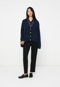 Femme portant un cardigan bleu marine boutonné sur une chemise bleue marine transparente, un pantalon noir, des mocassins noirs et portant un petit sac à bandoulière noir.