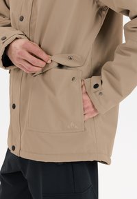 Beige vattentät jacka med tryckknappar, en framficka och en slät yta. Har en broderad logotyp på fickan.
