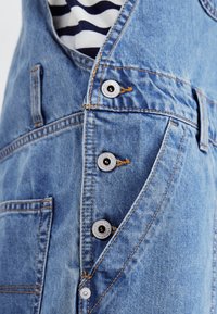 Salopette en denim de couleur bleu clair, avec des boutons-pression en métal argenté et des coutures orange contrastantes. Comprend une poche avant pour une fonctionnalité supplémentaire.