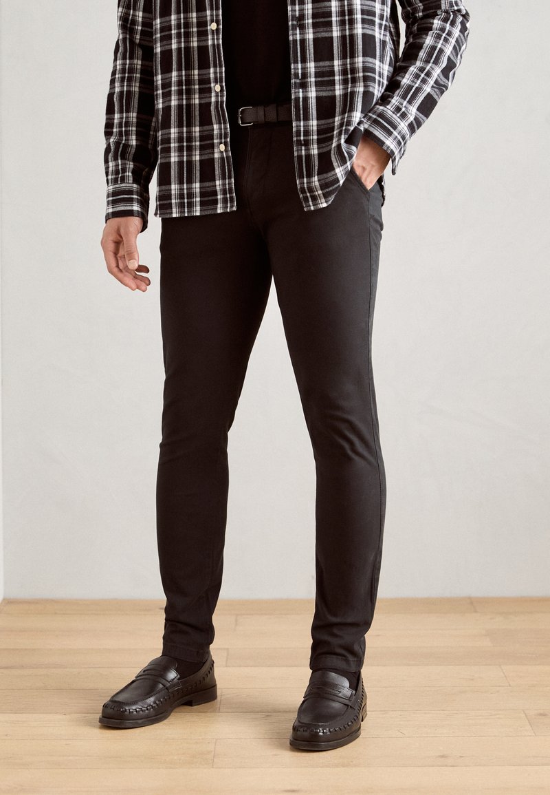 Pantaloni neri attillati abbinati a una maglietta nera, coperti da una camicia a maniche lunghe a scacchi bianca e nera. Scarpe in pelle nera completano il look.