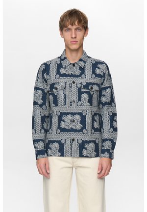 Jonge man draagt een donkerblauw overhemd met witte bloemmotieven en lichtbeige broek, staand tegen een effen witte achtergrond.