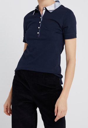 Osoba ubrana w granatowe polo z krótkim rękawem, z kołnierzykiem w kratę i plisą na guziki, połączone z czarnymi sztruksowymi spodniami na jednolitym tle.