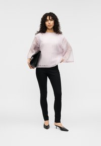Pull rose clair en maille avec manches amples en forme de cloche, associé à un pantalon noir ajusté et une pochette noire. Texture lisse et légèrement transparente.