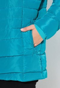 Ruka s jemně růžovým lakem na nehtech uvnitř kapesního zipové bundy v tyrkysové barvě, viditelný manžeta rukávu, na světlejším pozadí.