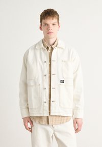 Cream denim jacket with two large front pockets, button closure, and contrasting stitching. Layered over a beige collared shirt.