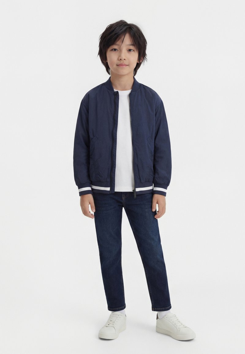 Νεαρός αγόρι στέκεται, φορώντας ναυτικό μπουφάν τύπου bomber, λευκό μπλουζάκι, σκούρο μπλε τζιν και λευκά αθλητικά παπούτσια μπροστά σε απλό φόντο.