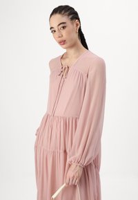 Svetloružová, voľná šaty s dlhými rukávmi, zúženým pásom a detailom so šnúrkou na výstrihu. Modelka drží malú bielu kabelku.