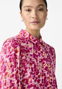 Collared blouse in vibrant pink, red, and orange abstract pattern. Soft fabric with subtle texture, featuring puffed sleeves and a buttoned front.