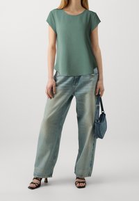 Top verde a maniche corte con scollatura rotonda, abbinato a jeans in denim a gamba larga di un blu chiaro e a una piccola borsa blu. Sandali neri con tacco.