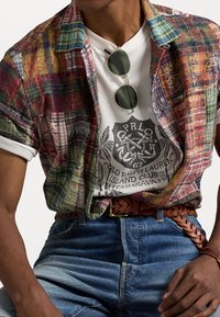 Wielokolorowa krótkozasięgowa koszula w kratę na białym t-shircie z grafiką, z okrągłymi ciemnymi okularami przeciwsłonecznymi, niebieskimi dżinsami i plecionym skórzanym paskiem.