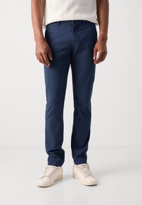 Pantalons bleu marine en tissu léger, avec une coupe droite, des poches avant et une fermeture éclair. Portés avec des baskets crème.