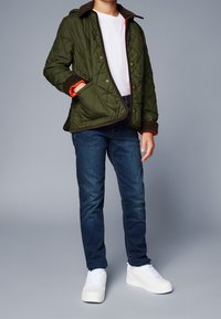 Tikkitakki vihreänä, ruskealla reunuksella ja oranssilla vuorella, yhdistetty tummiin farkkuihin ja valkoisiin lenkkareihin. Takissa on napitus ja taskut.