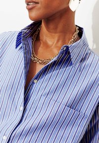 Chemise boutonnée bleu clair avec des rayures blanches et bleu marine, dotée d'une poche poitrine et d'un col structuré. Collier chaîne doré visible.