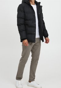 Sort pufferjakke med lynlås i front og hætte, med en quiltet design og en glat tekstur. Kombineret med lyse beige chinos og hvide sneakers.